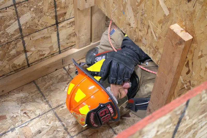 A firefighter's helmet and gloved hand push through a narrow opening in a wooden confined-space maze.