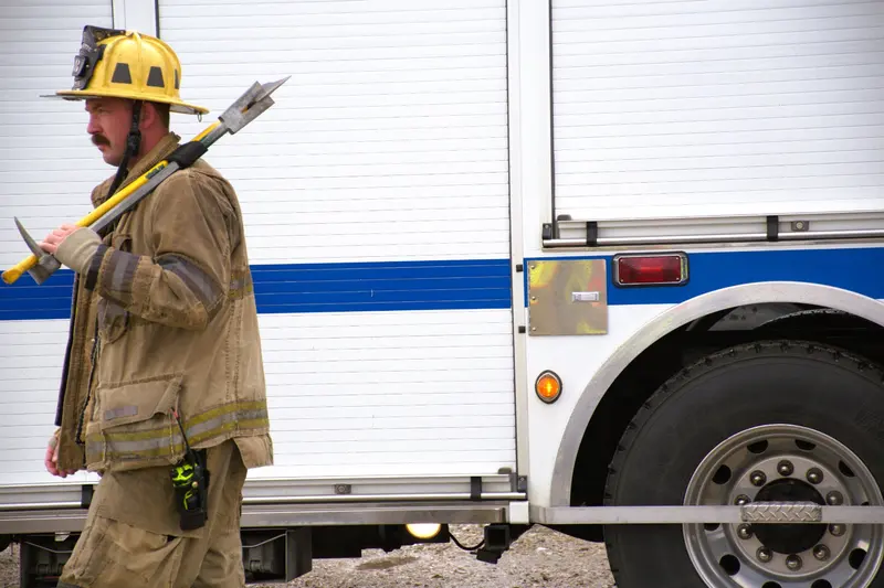 A firefighter carries a Halligan bar and an axe over their shoulder past a rescue truck.