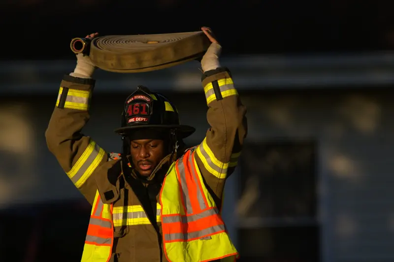 A firefighter in turnout gear and a hi-vis vest holds a rolled hose above their head.
