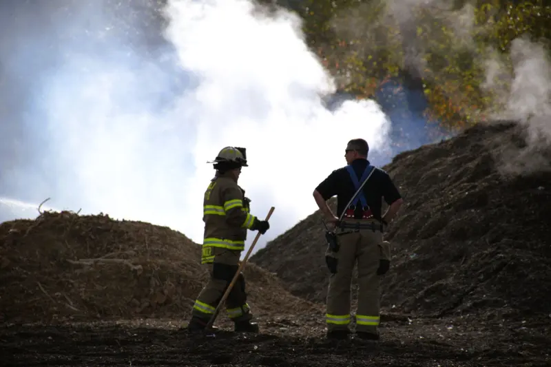 Two firefighters stand on a smoking mulch pile, silhouetted against white smoke.